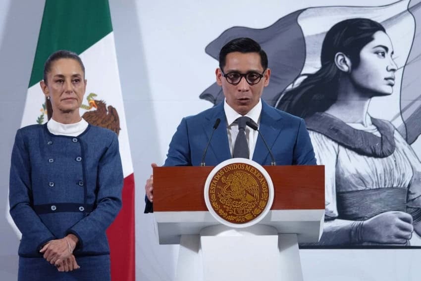 Mexican President Claudia Sheinbaum, left, at her daily press conference alongside journalist Miguel Angel Elorza, head of the website Infodemia. Both are onstage, Sheinbaum watching as Elorza speaks at a podium bearing the Mexican government's official seal. Elorza speaks to reporters off camera.
