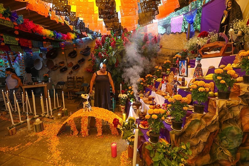 A woman stands next to a multi-tiered altar taller than she is, decked with photos, flowers and candles