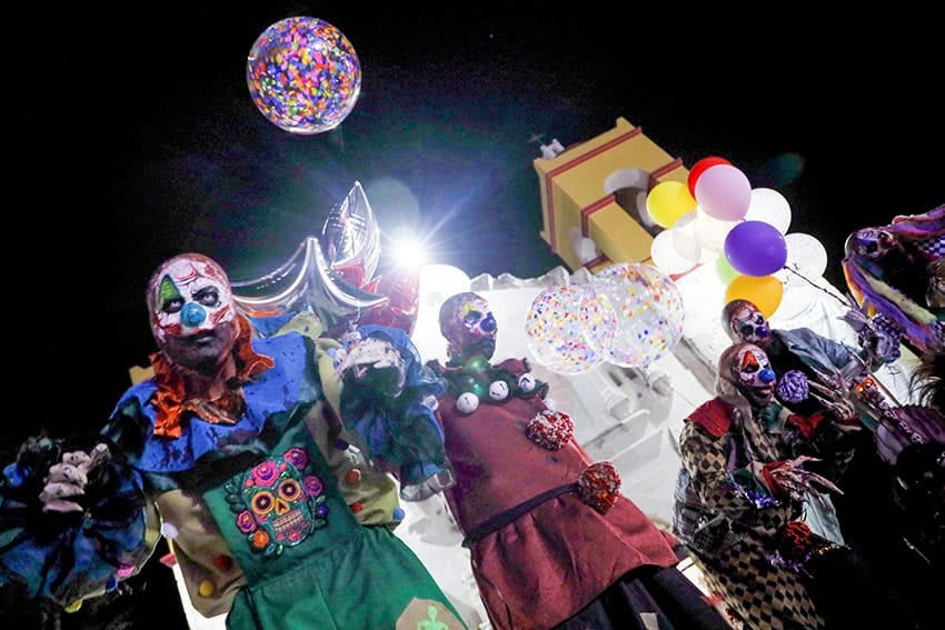 People in elaborate creepy clown costumes with references to Mexican catrinas and skulls stand in front of a church at night with balloons.