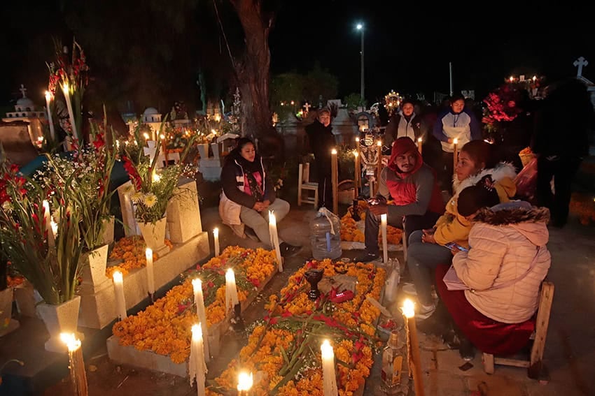 Warmly dressed families gather around graves lit with candles and covered in marigold flowers at night