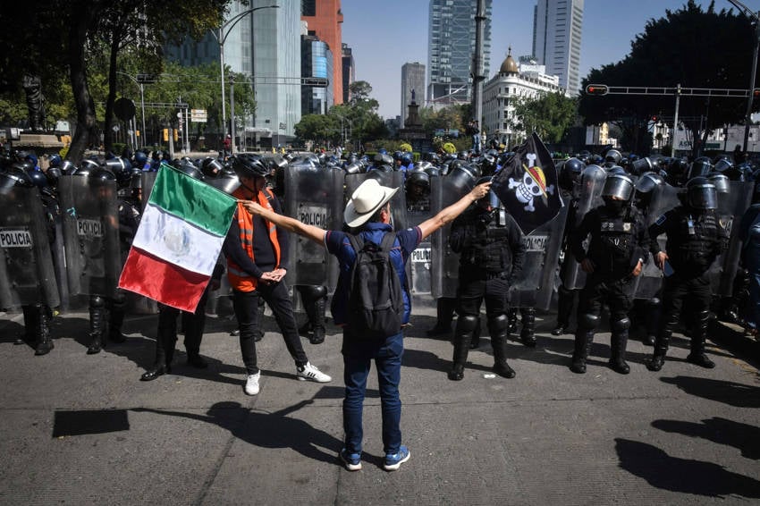 A lone protester wearing a cowboy hat and an orange vest stands with outstretched arms, holding a Mexican flag and another flag with a skull and crossbones, facing a line of Mexican riot police wearing helmets and holding shields on a sunny city street.