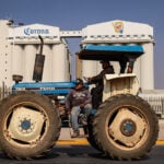 A farmer sits on a blue tractor in front of a Corona beer factory