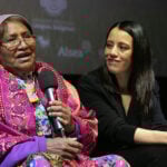 An older woman in colorful Indigenous clothing speaks into a microphone, next to a young Mexican woman dressed in black