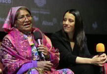 An older woman in colorful Indigenous clothing speaks into a microphone, next to a young Mexican woman dressed in black