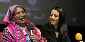 An older woman in colorful Indigenous clothing speaks into a microphone, next to a young Mexican woman dressed in black