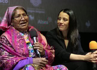 An older woman in colorful Indigenous clothing speaks into a microphone, next to a young Mexican woman dressed in black