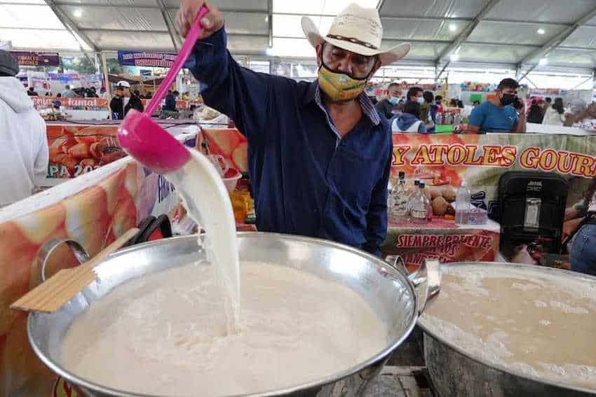 A Mexican food vendor in a cowboy hat and mask pours a ladleful of a thick, white traditional Mexican warm drink known as atole, into a large metal pot at a food festival.