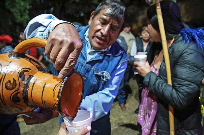 A Mexican food vendor in a cowboy hat and mask pours a ladleful of a thick, white traditional Mexican warm drink known as atole, into a large metal pot at a food festival.