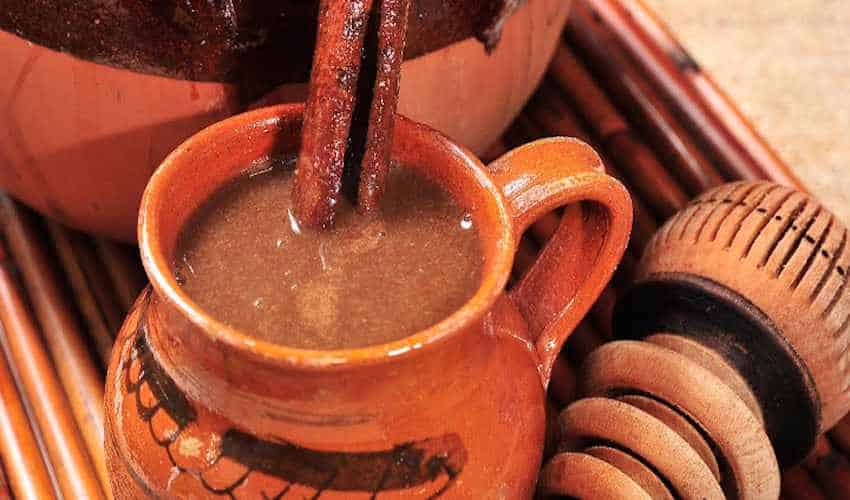 A mug of the thick, rich, traditional Mexican warm drink known as champurrado. It is served in an artisan clay mug with a cinnamon stick and stands next to to a Mexican wooden molinillo whisk.