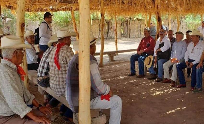 Local farmers in a rural area in Mexico wearing cowboy hats sit on benches under a thatched-roof shelter during an ejido assembly, illustrating the community governance process often required when buying land in Mexico to build a home in rural areas.