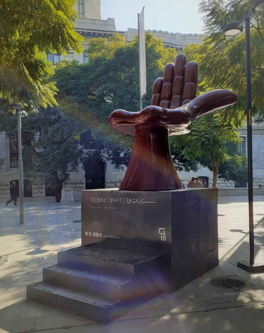 A monumental reddish-brown Hand Chair sculpture by Mexican artist Pedro Friedeberg sits atop a black stepped pedestal in the Alameda Central park of Mexico City, framed by green trees and historic architecture.