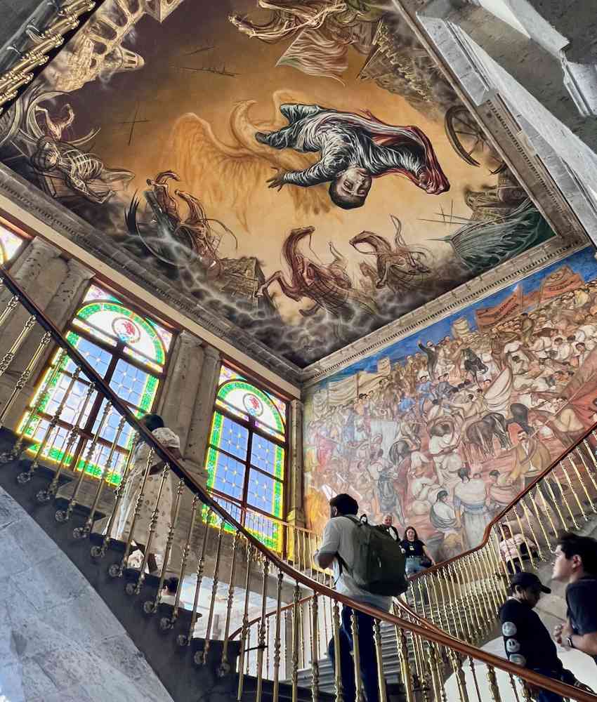 A wide, ornate stone staircase inside Chapultepec Castle shows visitors climbing the stairs, surrounded by a dramatic ceiling fresco depicting the conquest of Mexico by the Spanish, as well as mural on the wall beside the stairs.