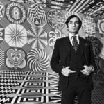 A black-and-white portrait of a young artist Pedro Friedeberg, wearing a dark suit and polka-dot tie, standing inside an immersive room covered entirely in geometric Op Art patterns and surrealist symbols.