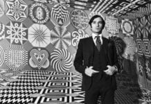 A black-and-white portrait of a young artist Pedro Friedeberg, wearing a dark suit and polka-dot tie, standing inside an immersive room covered entirely in geometric Op Art patterns and surrealist symbols.
