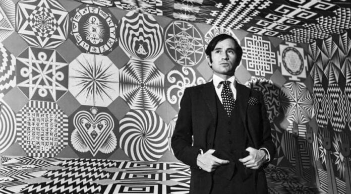 A black-and-white portrait of a young artist Pedro Friedeberg, wearing a dark suit and polka-dot tie, standing inside an immersive room covered entirely in geometric Op Art patterns and surrealist symbols.