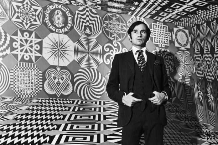 A black-and-white portrait of a young artist Pedro Friedeberg, wearing a dark suit and polka-dot tie, standing inside an immersive room covered entirely in geometric Op Art patterns and surrealist symbols.