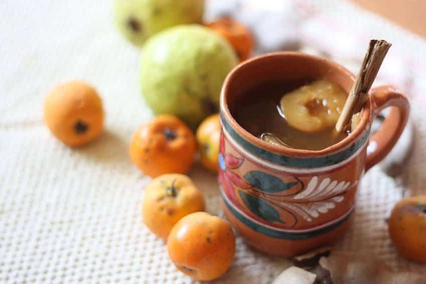 A colorful hand-painted clay mug (jarrito) filled with Ponche Navideño, one of Mexico's best-loved traditional Mexican warm drinks for Christmas, is garnished with a slice of fruit and a cinnamon stick, surrounded by tejocotes and guavas.