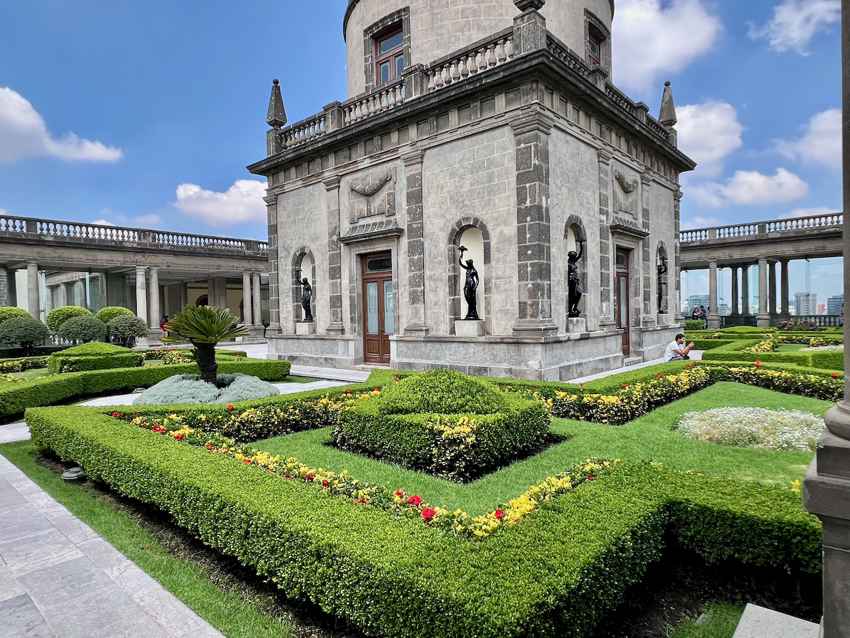 The beautifully manicured gardens on the rooftop maze of Chapultepec Castle, featuring symmetrical boxwood hedges, flower beds and a small cantera-brick building in the baroque style in the photo's center. The building is decorated with statues.