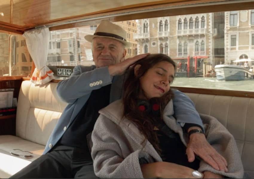 A candid shot of artist Pedro Friedeberg, wearing a beige fedora and light blue blazer, traveling via water taxi in Venice, Italy, while a female companion rests her head affectionately on his shoulder.