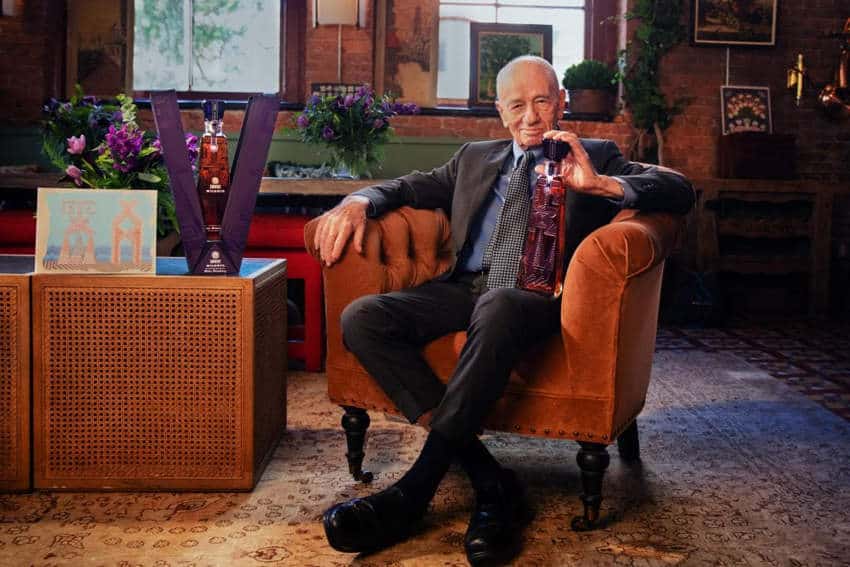 Mexican artist Pedro Friedeberg sits in a velvet armchair in an elegant room with antique brick walls, and an oriental rug on the floor. He is holding a limited edition bottle of tequila for Jose Cuervo that he designed with his signature surrealist artwork. The custom packaging for the bottle is displayed on a side table next to one of his artworks.