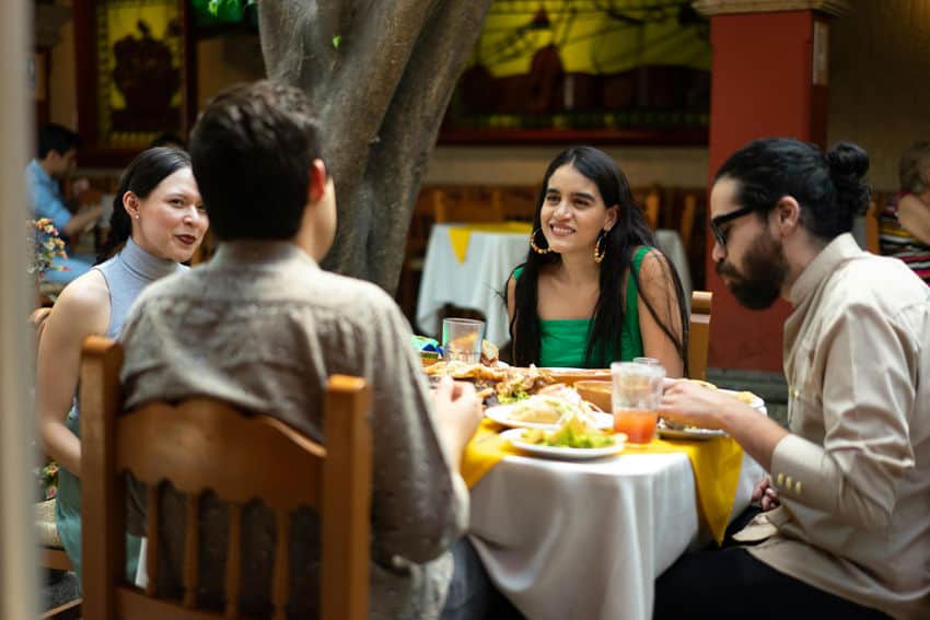 Two young men and two young women dine and converse around a restaurant table with Mexican food in a bright, tree-shaded courtyard restaurant.