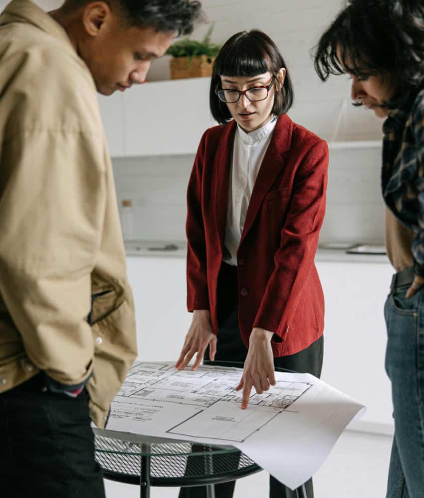 An architect in a red blazer reviews detailed floor plans with a male and female client, illustrating the design and planning phase for building a home in Mexico.
