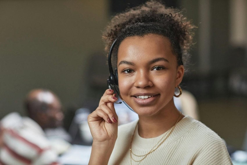 A smiling, professional young woman with curly hair wears a headset and holds the microphone near her mouth, representing the human element necessary when the limitations of AI language translation software are reached. She's wearing a cream-colored sweater and gold jewelry.