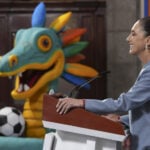 Sheinbaum at a press conference podium in front of a cartoon dragon or maybe quetzalcoatl with a soccer ball