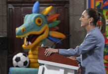 Sheinbaum at a press conference podium in front of a cartoon dragon or maybe quetzalcoatl with a soccer ball
