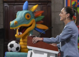 Sheinbaum at a press conference podium in front of a cartoon dragon or maybe quetzalcoatl with a soccer ball