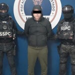 A detained man wearing a hoodie stands next to two uniformed SSPC agents.
