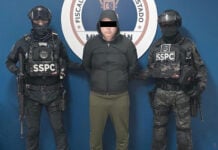 A detained man wearing a hoodie stands next to two uniformed SSPC agents.