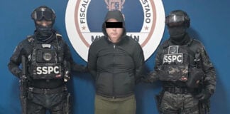 A detained man wearing a hoodie stands next to two uniformed SSPC agents.