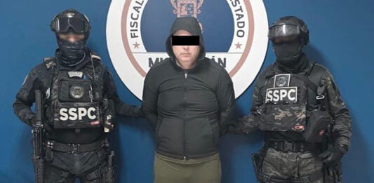 A detained man wearing a hoodie stands next to two uniformed SSPC agents.