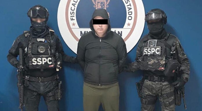 A detained man wearing a hoodie stands next to two uniformed SSPC agents.