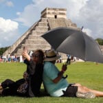 A woman takes a selfie while sitting back to back with a man holding a shade umbrella in front of a Chichén Itzá pyramid in Mexico