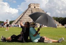 A woman takes a selfie while sitting back to back with a man holding a shade umbrella in front of a Chichén Itzá pyramid in Mexico