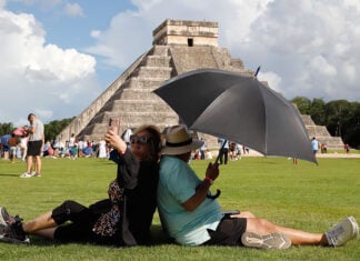 A woman takes a selfie while sitting back to back with a man holding a shade umbrella in front of a Chichén Itzá pyramid in Mexico
