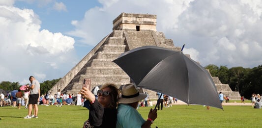 A woman takes a selfie while sitting back to back with a man holding a shade umbrella in front of a Chichén Itzá pyramid in Mexico