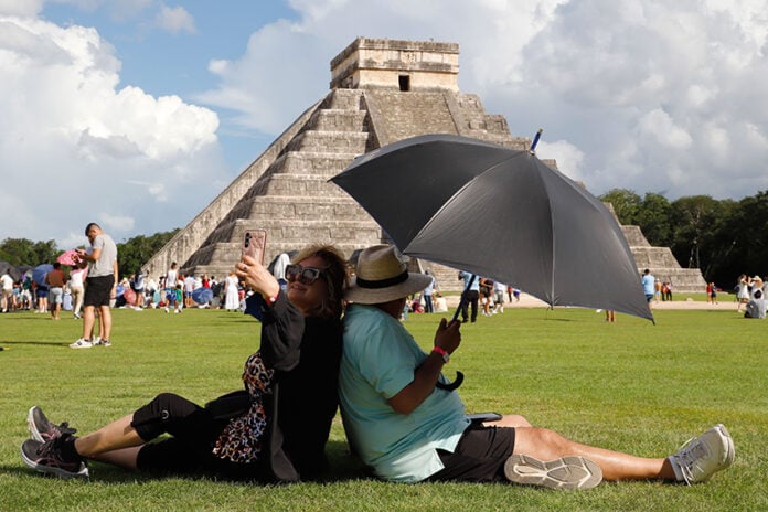 A woman takes a selfie while sitting back to back with a man holding a shade umbrella in front of a Chichén Itzá pyramid in Mexico