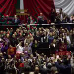 Ricardo Monreal stands at a podium in the Mexican Chamber of Deputies (congress chambers) surrounded by dozens of supporters with their fists raised in the air