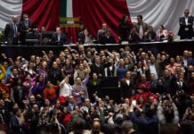 Ricardo Monreal stands at a podium in the Mexican Chamber of Deputies (congress chambers) surrounded by dozens of supporters with their fists raised in the air