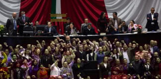 Ricardo Monreal stands at a podium in the Mexican Chamber of Deputies (congress chambers) surrounded by dozens of supporters with their fists raised in the air