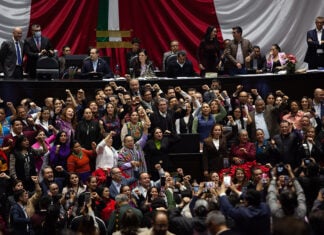 Ricardo Monreal stands at a podium in the Mexican Chamber of Deputies (congress chambers) surrounded by dozens of supporters with their fists raised in the air