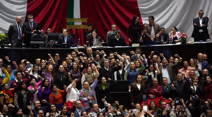 Ricardo Monreal stands at a podium in the Mexican Chamber of Deputies (congress chambers) surrounded by dozens of supporters with their fists raised in the air