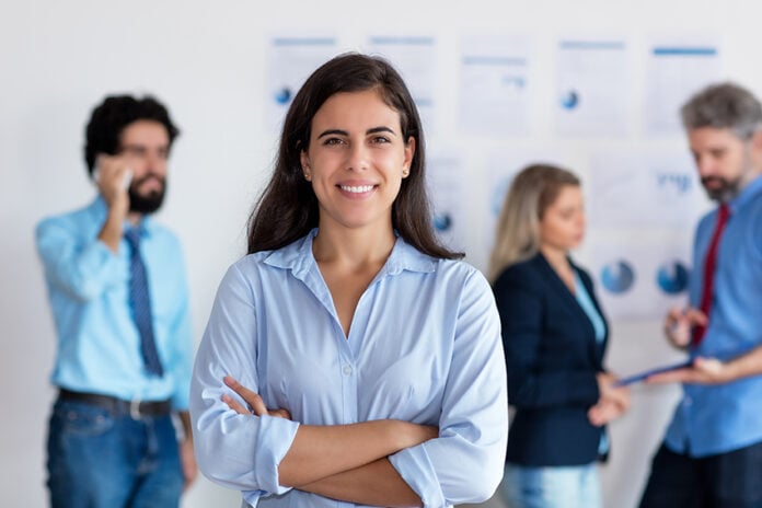 Mexican woman smiles at the camera in front of a group of coworkers all in business casual attire