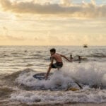 "A young boy skimboarding on a wave during a golden sunset, illustrating the active coastal lifestyle featured in Puerto Vallarta community news December 2025."
