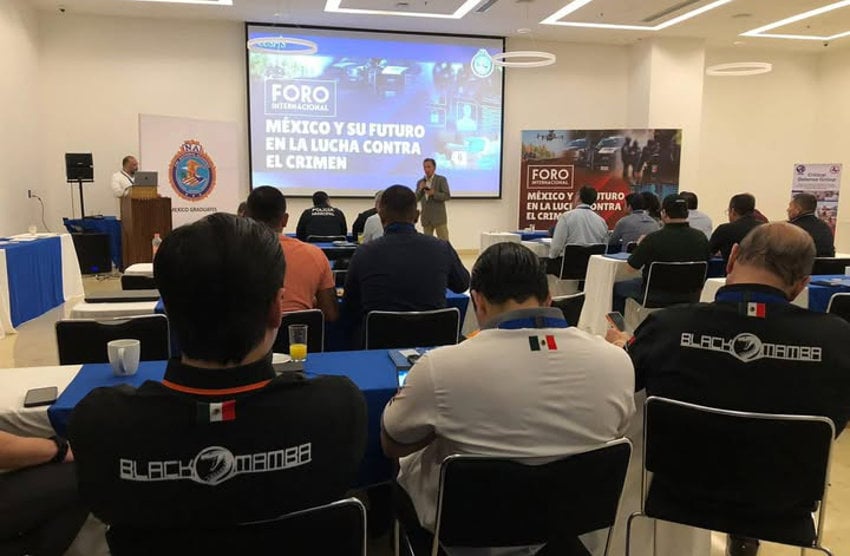 Attendees at the International Forum on Mexico and its Future in the Fight Against Crime, a security event in Puerto Vallarta in 2025, sit at conference tables in rows as a man in a suit speaks into a microphone.