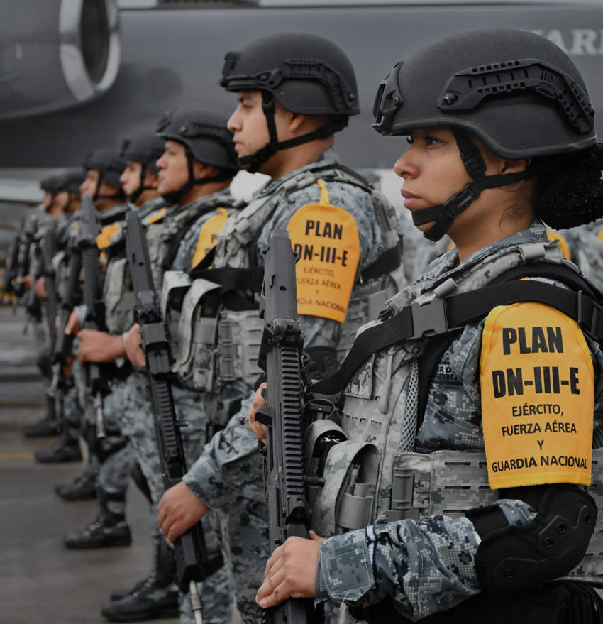 Members of the Mexican National Guard wearing Plan DN-III-E armbands standing in formation by a plane bearing machine guns and helmets on their heads.