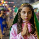 A young girl with hands clasped in prayer wears a pink and green costume evoking the traditional outfit of the Virgin of Guadalupe during a Fiestas Guadalupanas procession, a highlight of traditional Puerto Vallarta December 2025 events in the downtown district.